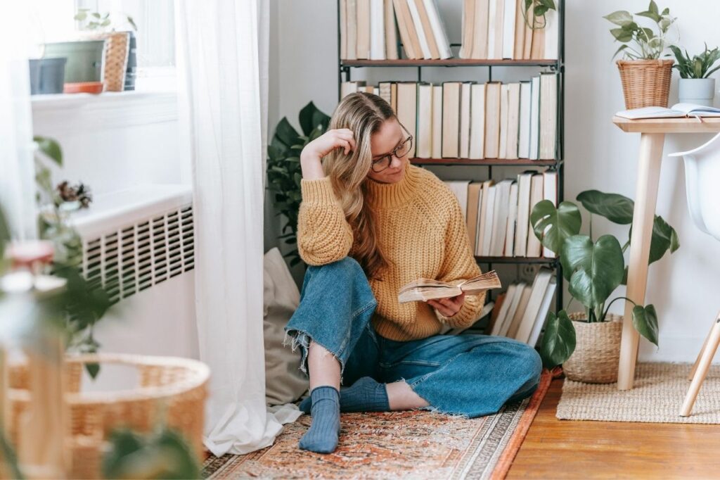 A woman sitting on the floor reading an English learning book near a bookshelf and plants, showing peaceful home study vibes.