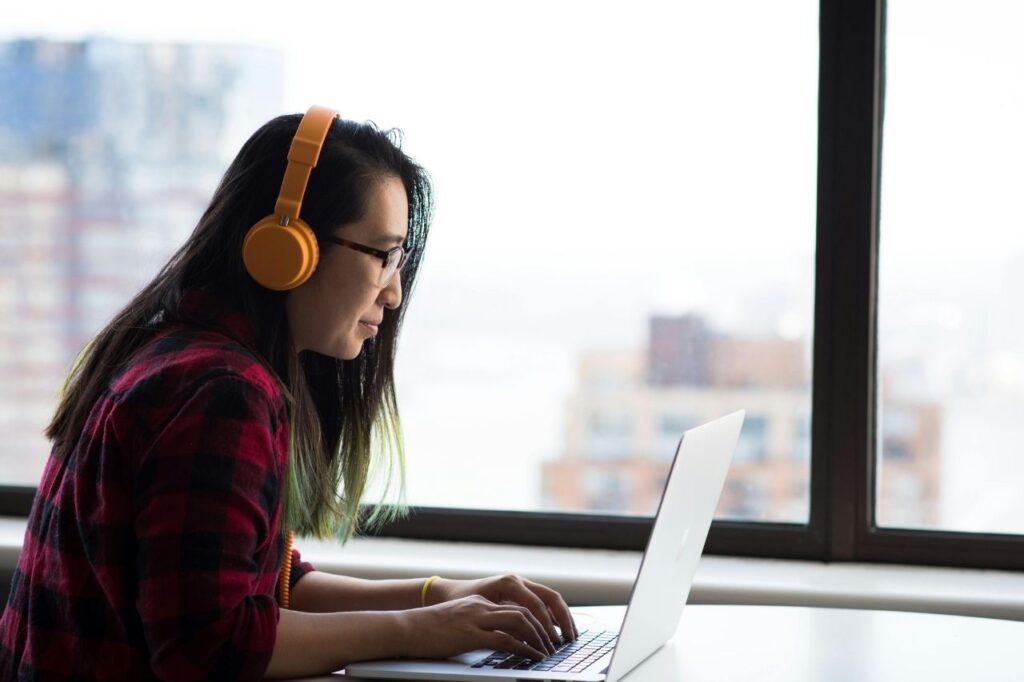 A young learner studying English on a laptop with headphones, sitting near a bright window.