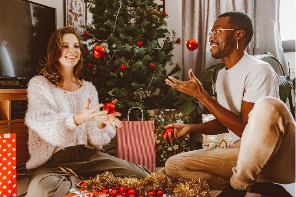 Two friends smiling and playing with red Christmas ornaments near a decorated tree.