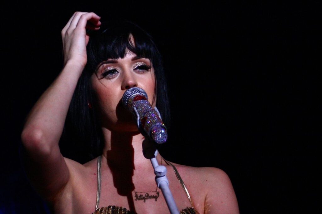 Katy Perry biography image showing her performing live with a sparkling microphone and emotional expression under stage lights