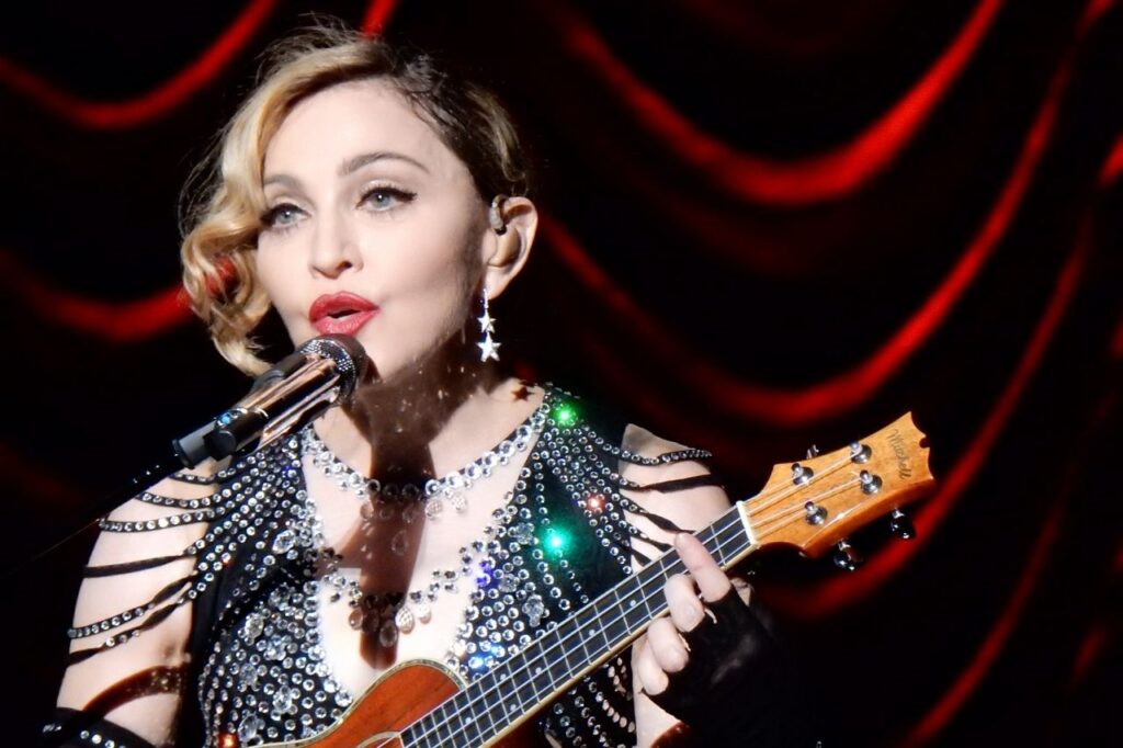 Madonna singing live on stage with her ukulele, surrounded by soft red stage lights